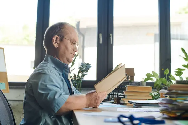 A man with inclusivity sits at his desk, engrossed in reading a book, surrounded by stacks of papers and books. — Stock Photo