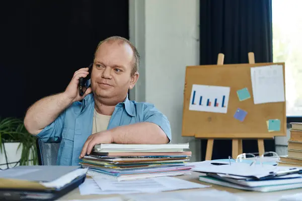 Man with inclusivity at desk with phone, surrounded by paperwork in busy office. — Stock Photo