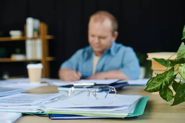 A pair of glasses sits on top of a stack of papers on a desk, with a man with inclusivity working in the background. — Stock Photo