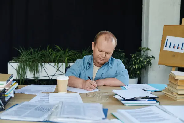Ein Mann mit Inklusivität sitzt an einem Schreibtisch und schreibt auf ein Blatt Papier, umgeben von Papierkram und einer Tasse Kaffee.. — Stockfoto