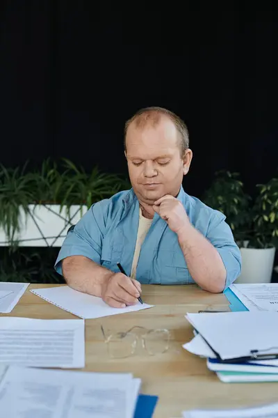 Ein Mann mit Inklusivität sitzt an einem Schreibtisch und schreibt auf ein Blatt Papier, umgeben von anderen Papieren. — Stockfoto