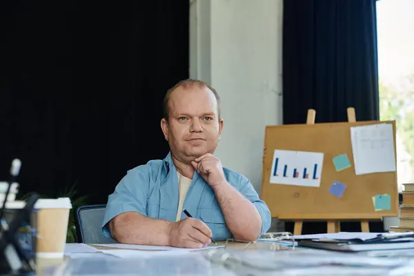 Ein inklusiver Mann sitzt an einem Schreibtisch und arbeitet nachdenklich an einem Dokument, während er direkt in die Kamera blickt.. — Stockfoto