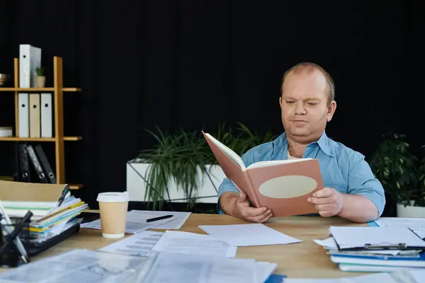 Ein inklusiver Mann sitzt an seinem Schreibtisch, umgeben von Papieren, und liest ein Buch. — Stockfoto