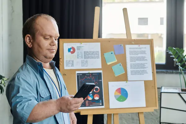 A man with inclusivity looks at his phone while standing near a bulletin board with graphs and notes. — Stock Photo
