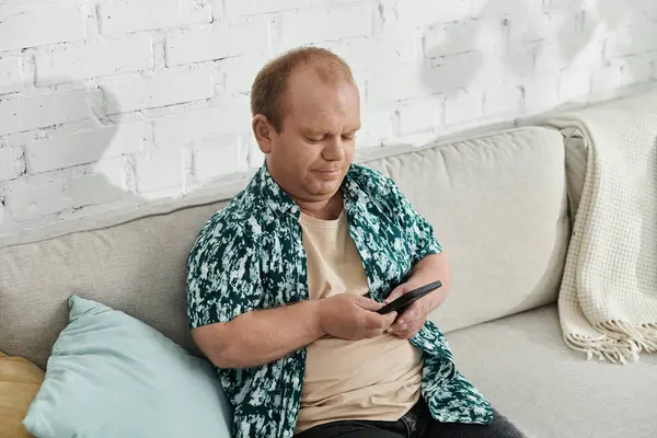 A man with inclusivity in a patterned shirt sits on a couch, looking down at his phone. — Stock Photo