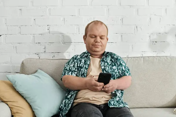 A man with inclusivity sits on a couch with a white brick wall background, looking at his smartphone. — Stock Photo