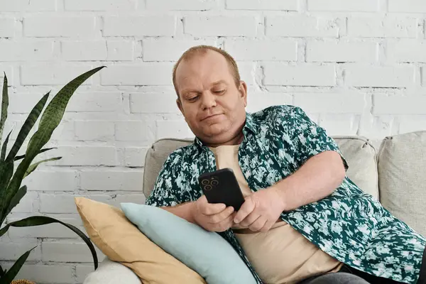 A man with inclusivity in a patterned shirt sits on a sofa, using his smartphone. — Stock Photo