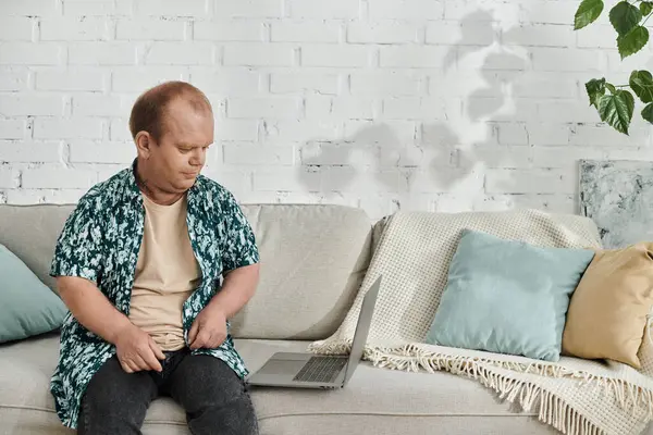 A man with inclusivity sits on a couch, adjusting his shirt, with a laptop on the sofa beside him. — Stock Photo