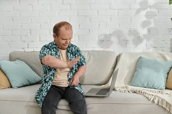 A man with inclusivity sits on a couch, gesturing with his right hand while looking at a laptop. — Stock Photo