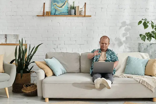 Un uomo con inclusività è seduto su un divano in un ambiente home office, lavorando diligentemente sul suo computer portatile. — Foto stock
