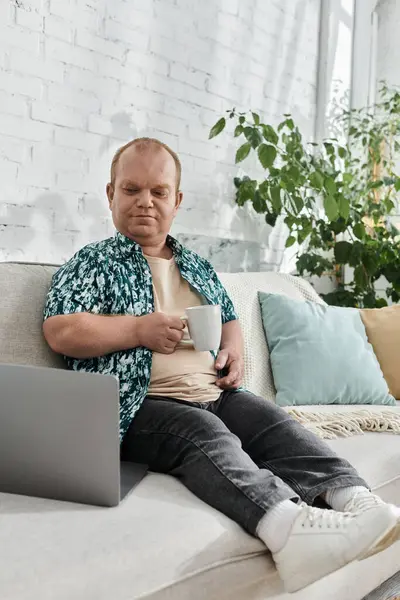 A man with inclusivity sits on a white couch, enjoying a cup of coffee and working on his laptop. — Stock Photo