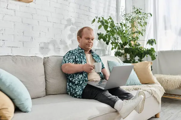 A man with inclusivity sits comfortably on a couch, enjoying a cup of coffee and working on his laptop. — Stock Photo