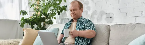 A man with inclusivity sits on a couch, working on a laptop while holding a mug of coffee. — Stock Photo
