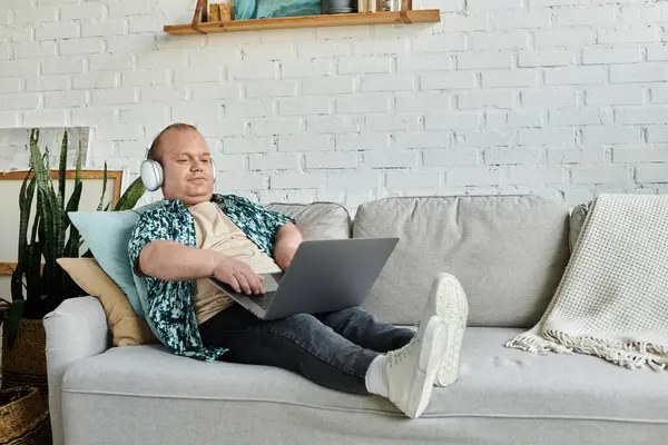 Ein Mann mit Inklusivität sitzt auf einer Couch und arbeitet an einem Laptop, während er Kopfhörer trägt. — Stockfoto