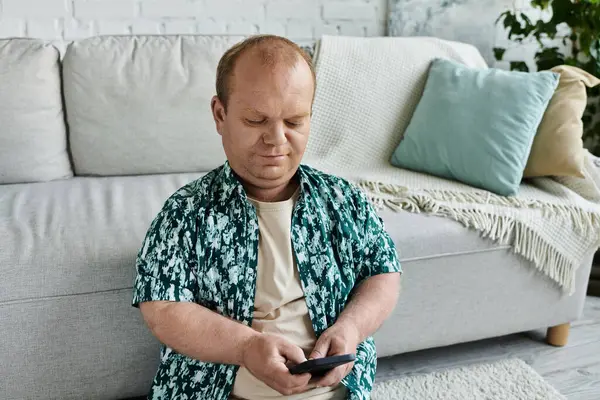 A man with inclusivity in a patterned shirt sits on the floor in front of a sofa, using his smartphone. — Stock Photo