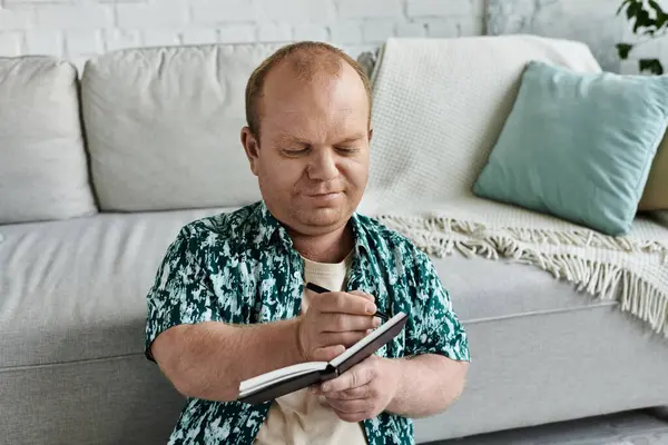 Ein inklusiver Mann sitzt auf dem Boden neben einer Couch und konzentriert sich darauf, in ein Notizbuch zu schreiben.. — Stockfoto