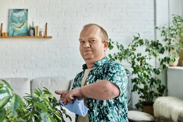Un homme avec inclusivité dans une chemise verte tend à des plantes dans une maison avec un mur de briques blanches. — Photo de stock