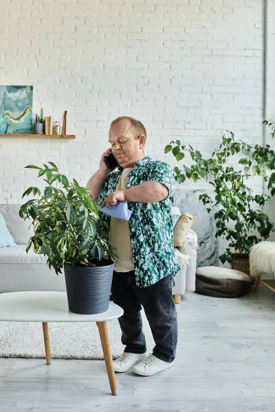 Um homem com inclusividade falando em seu telefone enquanto cuida de sua planta interior. — Fotografia de Stock