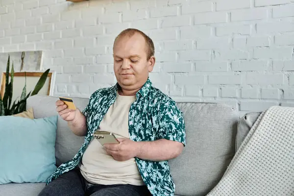 A man with inclusivity sits on a couch, using his phone and holding a credit card. — Stock Photo