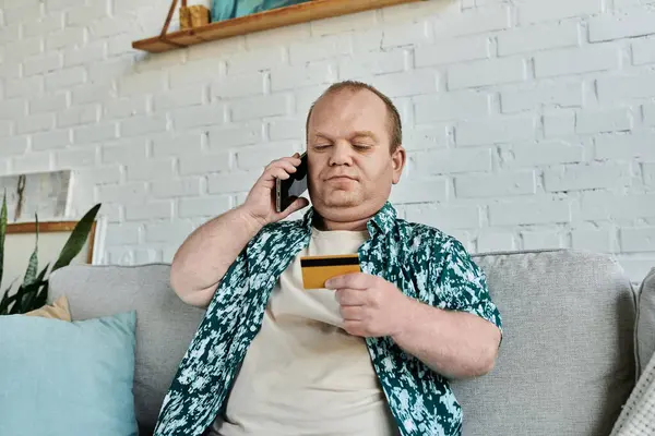 A man with inclusivity sits on a couch, talking on his phone and holding a credit card. — Stock Photo