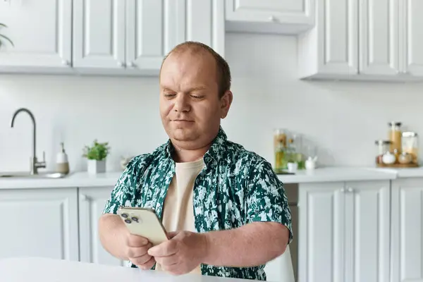 A man with inclusivity wearing a patterned shirt sits at a kitchen table, intently looking at his smartphone. — Stock Photo