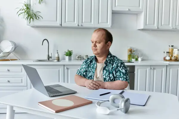 A man with inclusivity sits at a table in a kitchen, working on a laptop. — Stock Photo