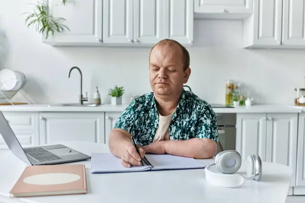 Ein Mann im grünen Hemd schreibt in ein Notizbuch an einem Tisch in einer weißen Küche.. — Stockfoto
