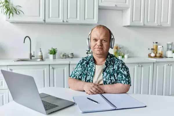 Um homem com inclusividade usando fones de ouvido se senta em uma mesa de cozinha trabalhando em um laptop e notebook. — Fotografia de Stock