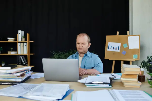 Um homem com inclusividade senta-se em uma mesa em um escritório desordenado, focado em seu laptop. — Fotografia de Stock
