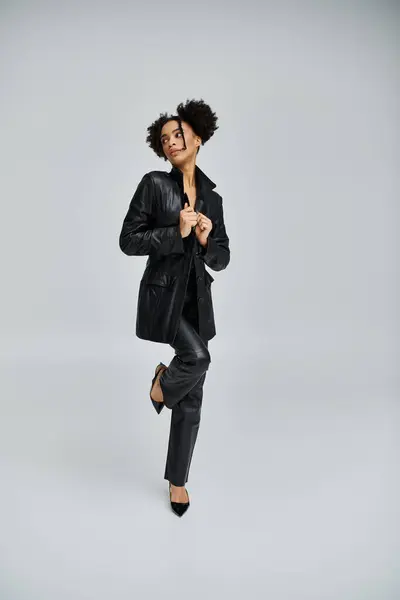A stylish young woman in a black leather jacket and pants poses against a gray background, exuding confidence and trendsetting fashion. — Stock Photo