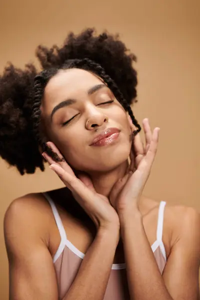Mulher negra jovem sorri com os olhos fechados, mãos no rosto, em tranças e top de tanque, contra fundo bege. — Fotografia de Stock