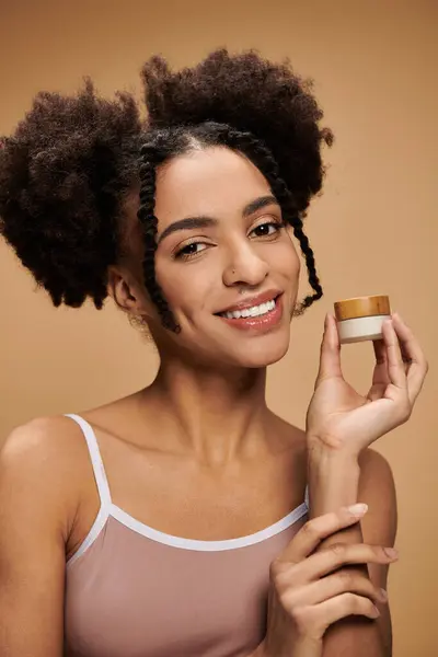 Una joven afroamericana con una sonrisa brillante sostiene un frasco de crema en su mano sobre un fondo beige. — Stock Photo