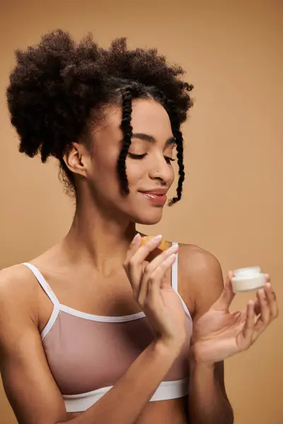 A young African American woman holds a cream jar in front of a beige background. — Stock Photo