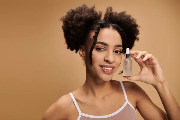A young African American woman smiles while holding a bottle of serum against a beige background. — Stock Photo