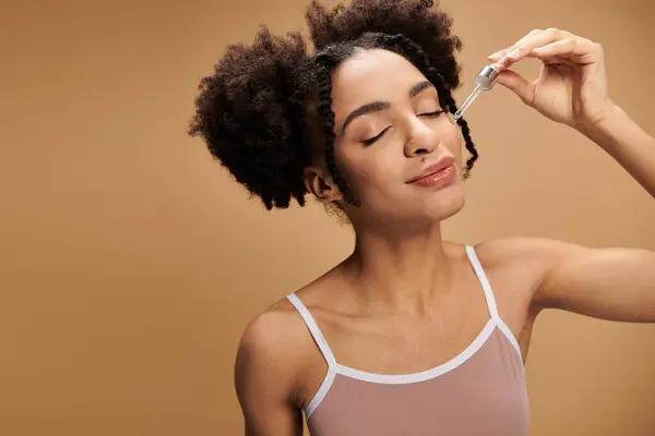 Uma jovem afro-americana com pele bonita aplica soro ao rosto, mostrando um brilho radiante contra um fundo bege.. — Fotografia de Stock