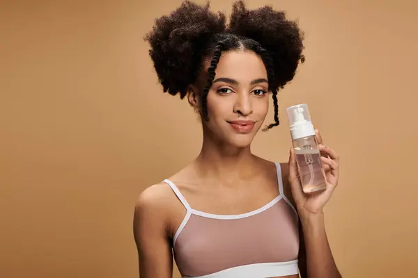 A young African American woman with beautiful skin and a radiant smile holds a bottle of skincare product against a warm beige background. — Stock Photo