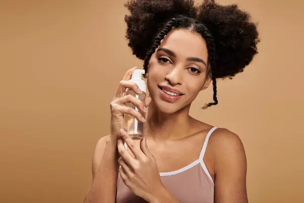 A young African American woman with a warm smile holds a bottle of skincare product against a beige background. — Stock Photo