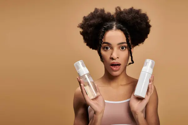 Young Black woman holds two bottles, looking surprised and slightly concerned against a beige background. — Stock Photo