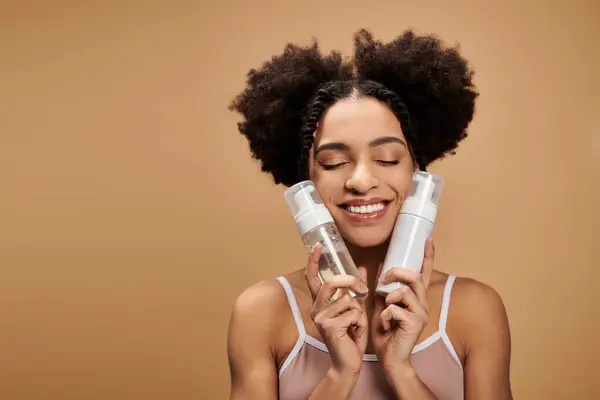 A young African American woman with a beautiful smile holds two bottles of skincare products against her face. — Stock Photo