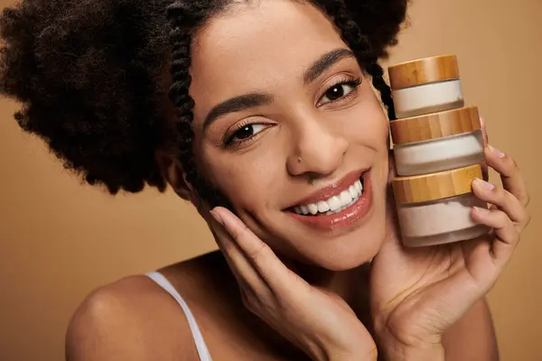 A young African American woman with a radiant smile holds skincare products against her face. — Stock Photo