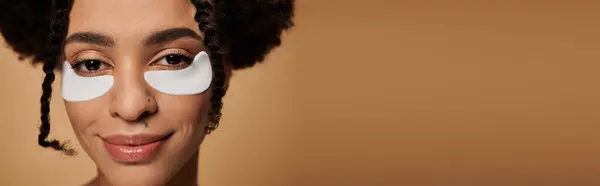 A close-up of a young African American woman with under-eye patches, smiling gently against a beige background. — Stock Photo