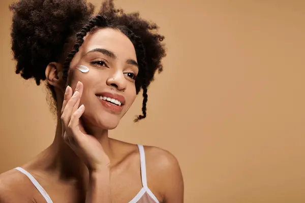 A young African American woman with a warm smile applies moisturizer to her face, showcasing the beauty of diversity. — Stock Photo