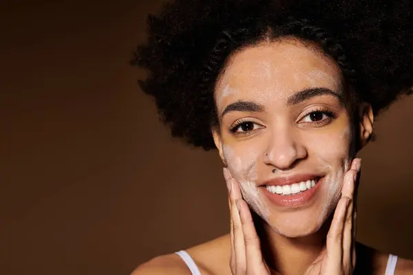 A young African American woman smiles brightly as she applies a face mask, highlighting the beauty and diversity of skin care practices. — Stock Photo