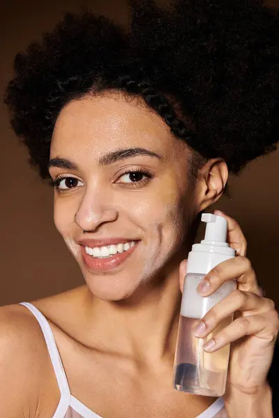 A young African American woman smiles while holding a bottle of facial cleanser. — Stock Photo