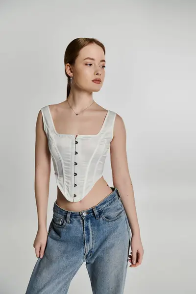 A young woman stands against a white backdrop, wearing a white corset and blue jeans. — Stock Photo