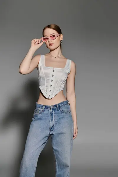 A young woman poses against a gray backdrop, wearing a white corset top and blue jeans. — Stock Photo