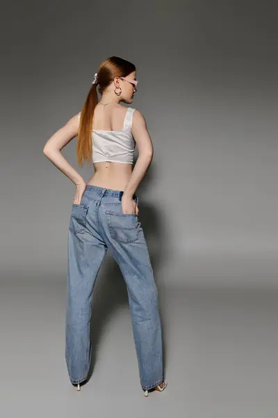 Una giovane donna in jeans denim e un crop top bianco posa su uno sfondo grigio. — Foto stock