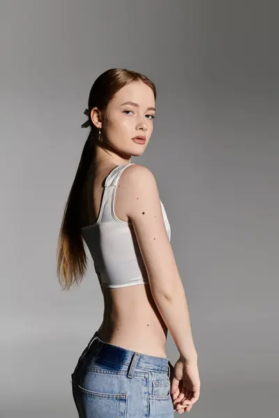 Una giovane donna posa in un top bianco e jeans su uno sfondo grigio. — Foto stock