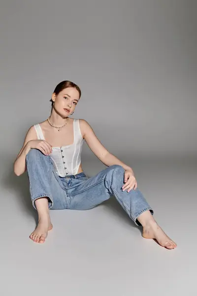 Une jeune femme pose en toute confiance dans un haut corset blanc et un jean bleu sur fond gris. — Photo de stock