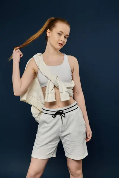 Giovane donna con i capelli rossi in coda di cavallo, canotta bianca e pantaloncini posa sullo sfondo blu. — Foto stock
